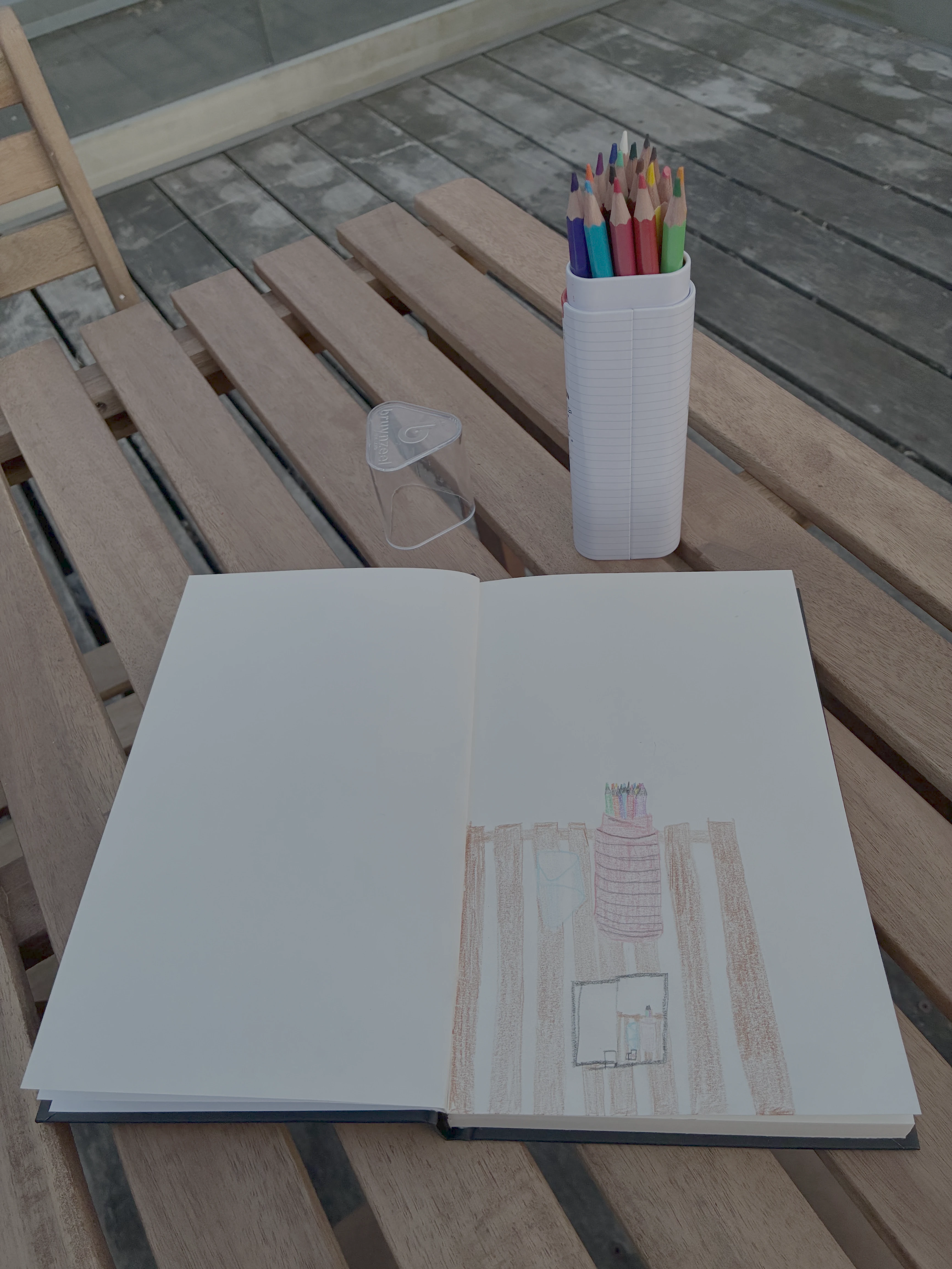  a picture of a table with a tall jar of colored pencils and the cap laying next to it. underneath is a sketchbook, which has a drawing of the pencils themselves. inside the drawing is another sketchbook, with a tiny miniature nested pencil drawing. 