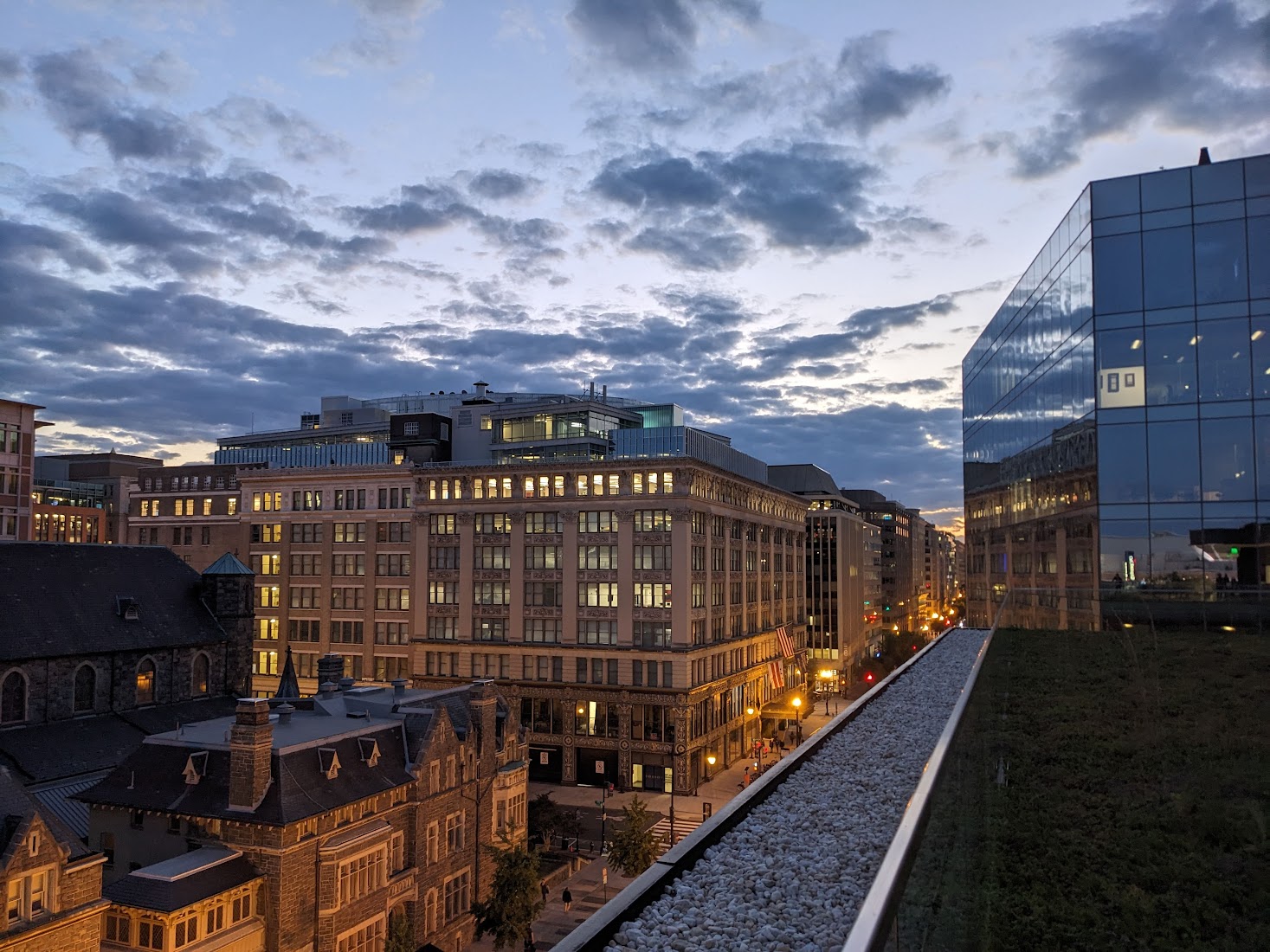 a picture of a city, taken from a tall rooftop. there is a glass building to the right and ornate architectures to the left. the street is filled with lights. far above, the clouds are dark grey against a bright setting sun.