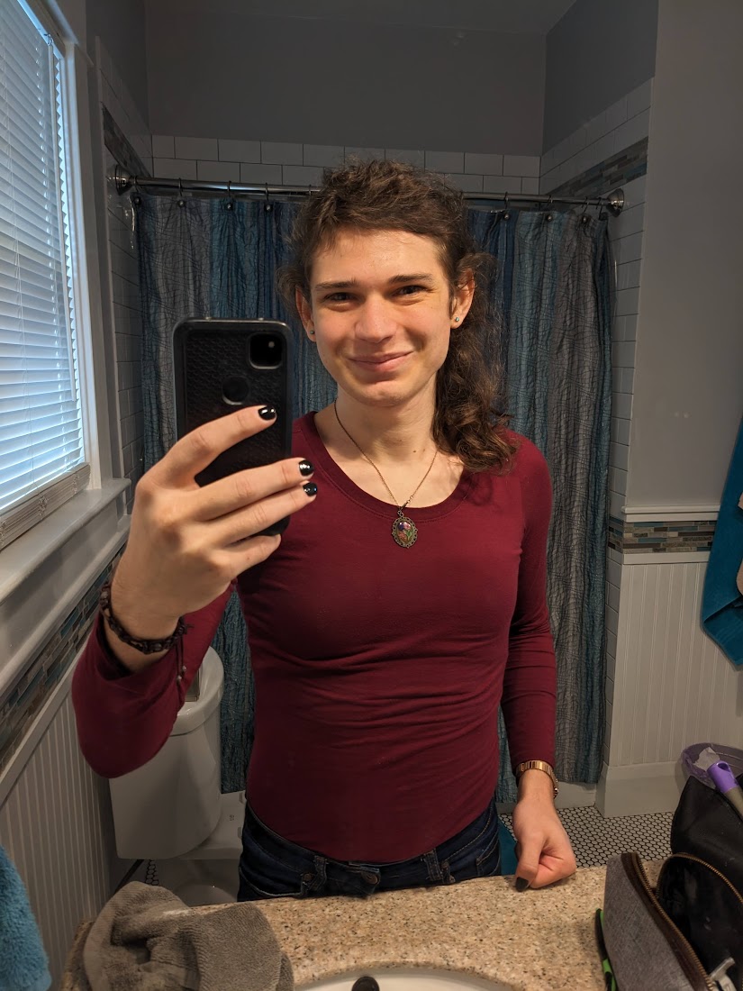 mirror selfie!! i'm wearing a long sleeved burgundy shirt that hugs my curves and jeans that are just visible at the waist. my nails are painted glossy black. i'm wearing a bronze-ish metal watch, a brown bracelet, and a delicate necklace with a picture of a flower. my hair is half up and drapes over my left shoulder. i'm smiling wide, bringing out my laugh lines, although my mouth is closed.